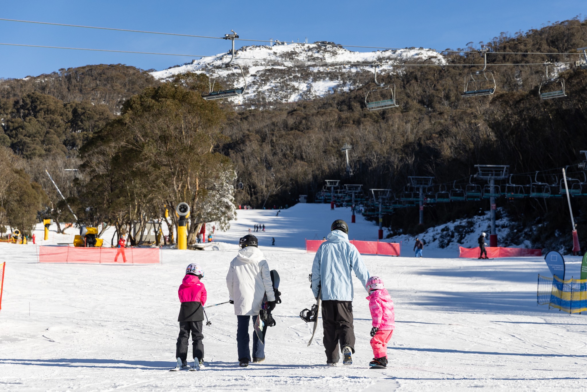 A family walking onto a groomed Australian ski slope with snowmaking equipment visible in the background